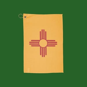 Puede incluir: Una toalla de golf amarilla con un símbolo Zia rojo, un símbolo solar nativo americano, centrado en la toalla. La toalla tiene una arandela metálica y un lazo para colgar. El fondo es verde liso.