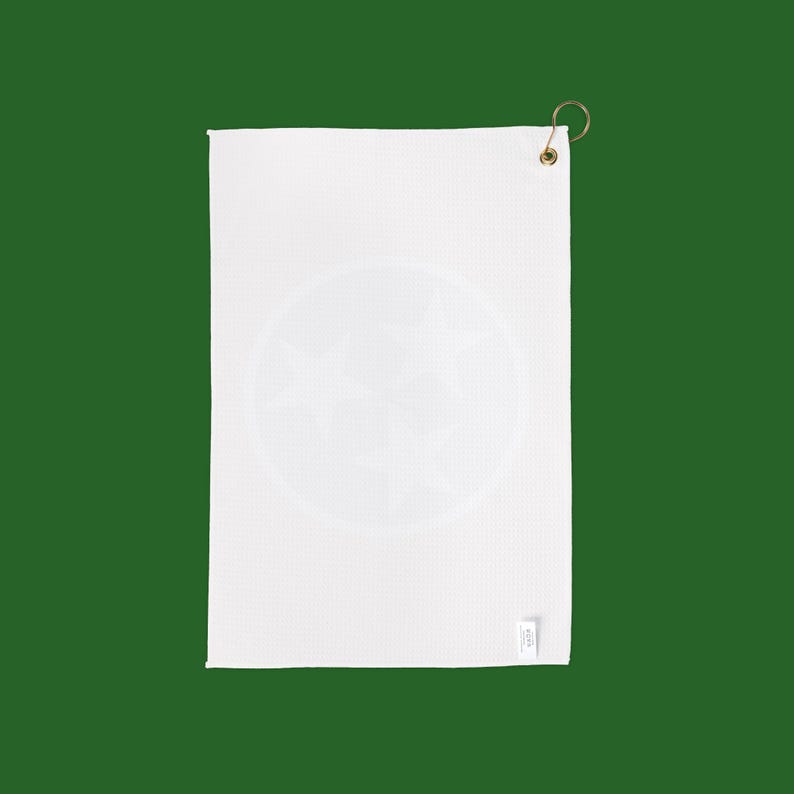 May include: White golf towel with a circular design featuring three stars. The towel has a textured surface and a brass-colored hook for hanging. A small tag is attached to the bottom right corner.