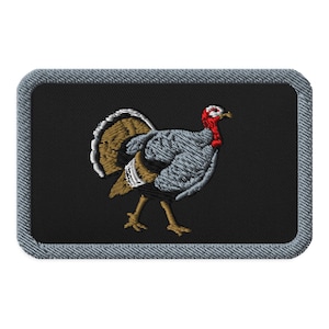 May include: A rectangular embroidered patch featuring a turkey with grey and brown feathers, a bright red neck, and a white head. The design is set against a solid black background and is framed by a thick, textured grey border with rounded corners.