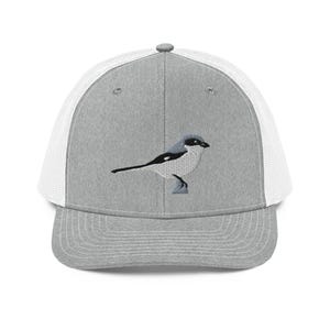 Puede incluir: Gorra de camionero gris con parte trasera de malla blanca y visera curva. La gorra presenta un diseño bordado de un pájaro en negro, blanco y gris. El pájaro está posado en una pequeña rama.