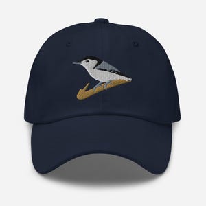 Puede incluir: Una gorra de béisbol azul marino con un bordado de un trepador de pecho blanco con plumaje negro, blanco y gris posado sobre una rama marrón. La gorra tiene una corona estructurada, una visera curva y un botón en la parte superior central.