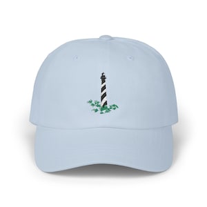 May include: A light blue baseball cap with an embroidered black and white lighthouse with green foliage.