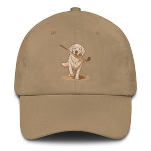Könnte beinhalten: Beige Baseball Cap mit einer Illustration eines Golden Retrievers, der einen Golfschläger im Maul hält. Der Hund steht auf einem Sandfleck. Die Kappe hat einen gebogenen Schirm und einen verstellbaren Riemen.