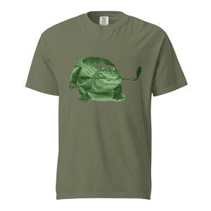 May include: An olive green t-shirt with a green illustration of an alligator holding a golf club. The alligator has detailed scales and is on a patch of green grass. The t-shirt is a casual wear item, suitable for everyday use.