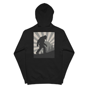 May include: Black hooded sweatshirt featuring a Bigfoot silhouette walking across a mountain landscape with a sunburst design. The graphic is in shades of gray and white. The sweatshirt is a casual garment.