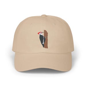 Puede incluir: Una gorra de béisbol beige con una imagen bordada de un pájaro carpintero de cabeza roja picoteando un tronco de árbol.