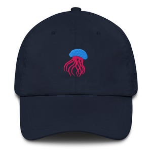 Könnte beinhalten: Marineblaue Baseballkappe mit einem gestickten Quallen-Design in Blau und Pink. Die Kappe hat einen gebogenen Schirm und einen verstellbaren Riemen. Das Quallen-Design ist ein lebendiger Kontrast zum dunklen Stoff.