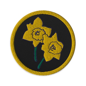 May include: A round, yellow embroidered patch with a black background featuring two yellow daffodils with green stems.