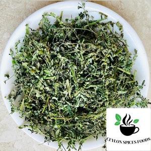 May include: A white bowl filled with dried green moringa leaves. The leaves are a light green color and have a slightly brown tint. The bowl is sitting on a white surface. The text "CEYLON SPICES FOODS" is visible in the bottom right corner of the image.