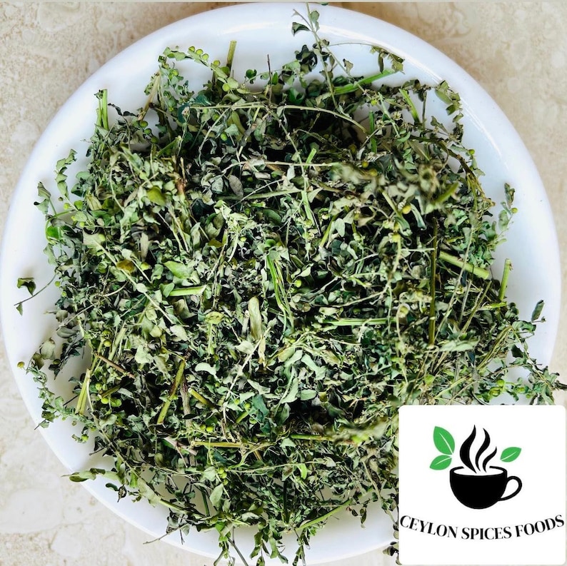 May include: A white bowl filled with dried green moringa leaves. The leaves are a light green color and have a slightly brown tint. The bowl is sitting on a white surface. The text "CEYLON SPICES FOODS" is visible in the bottom right corner of the image.