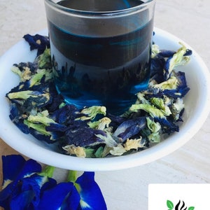 May include: A glass of blue butterfly pea flower tea with dried butterfly pea flowers in a white bowl. The tea is a vibrant blue color and the flowers are a mix of blue, green, and white.