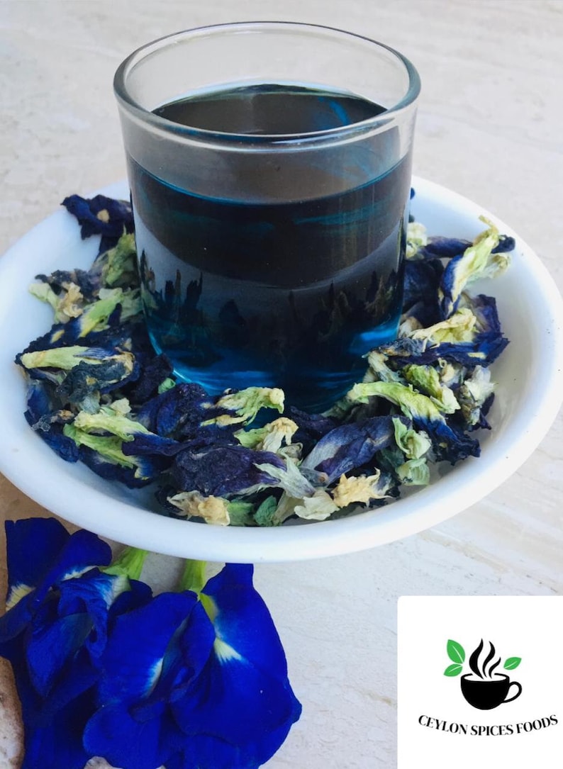 May include: A glass of blue butterfly pea flower tea with dried butterfly pea flowers in a white bowl. The tea is a vibrant blue color and the flowers are a mix of blue, green, and white.