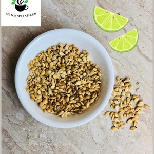 Puede incluir: Un cuenco blanco lleno de semillas de color marr&oacute;n claro, con un peque&ntilde;o mont&oacute;n de semillas al lado. Dos gajos de lima est&aacute;n en el fondo. El texto "CEYLON SPICES FOODS" est&aacute; en la esquina superior izquierda.