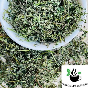 May include: A white bowl filled with dried green moringa leaves. The bowl is sitting on a white surface with more moringa leaves scattered around it.
