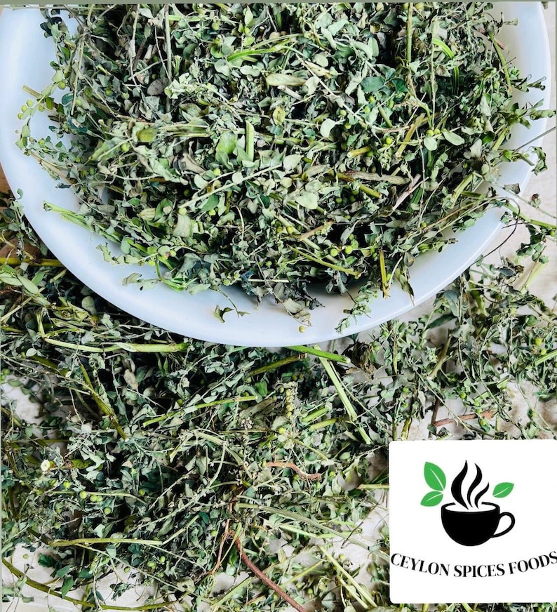 May include: A white bowl filled with dried green moringa leaves. The bowl is sitting on a white surface with more moringa leaves scattered around it.