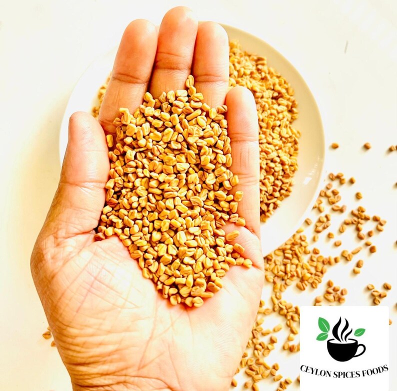Puede incluir: Una mano sosteniendo un mont&oacute;n de semillas de alholva de color marr&oacute;n dorado, con m&aacute;s semillas derram&aacute;ndose sobre una superficie blanca. Un plato blanco lleno de semillas est&aacute; en el fondo. El logotipo de Ceylon Spices Foods est&aacute; en la esquina inferior derecha.