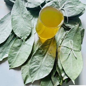 Può includere: Un bicchiere riempito con un liquido giallo vibrante circondato da diversi mazzetti di foglie essiccate verde pallido legate con spago bianco. Un logo nell'angolo presenta un'icona di tazza nera con foglie verdi e il testo CEYLON SPICES FOODS.
