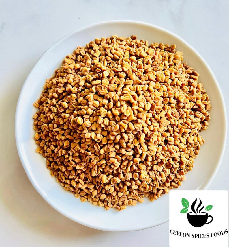 Puede incluir: Un plato blanco rebosante de semillas de alholva de color marr&oacute;n dorado. Las semillas son peque&ntilde;as, de forma irregular y densamente empaquetadas. El logotipo de Ceylon Spices Foods est&aacute; en la esquina inferior derecha.