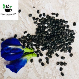 May include: A close-up of a pile of black butterfly pea flower seeds on a light brown surface. A blue butterfly pea flower is in the lower left corner of the image.