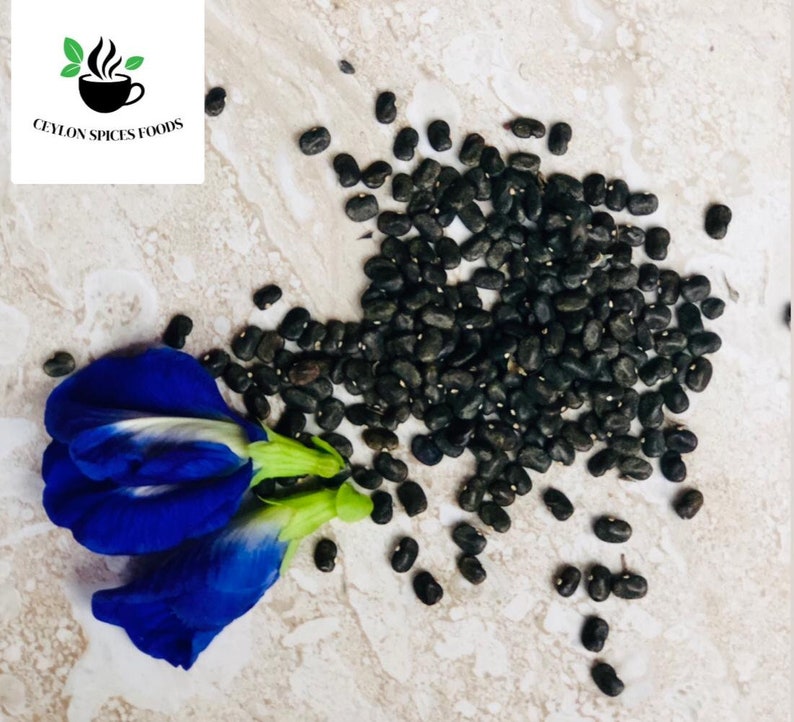 May include: A close-up of a pile of black butterfly pea flower seeds on a light brown surface. A blue butterfly pea flower is in the lower left corner of the image.