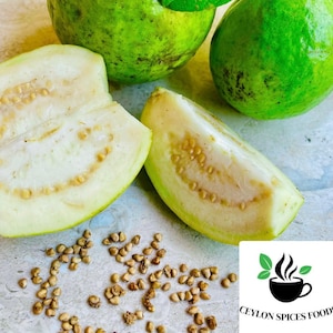 May include: Close-up of a white guava fruit cut in half, revealing the seeds inside. Two whole green guavas are also visible, along with scattered seeds on a light brown surface. The image includes the text "Ceylon Spices Foods".
