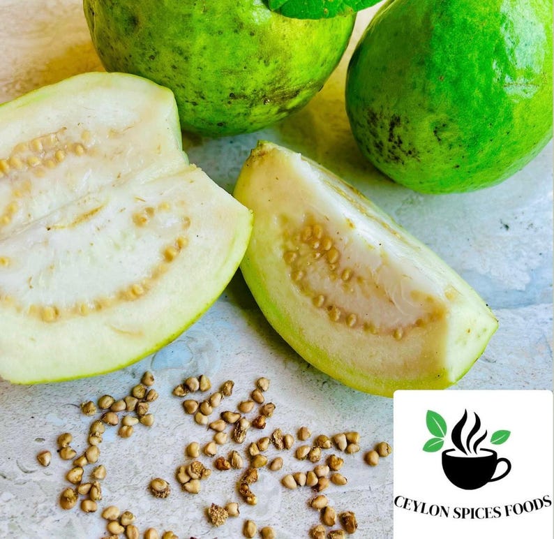 May include: Close-up of a white guava fruit cut in half, revealing the seeds inside. Two whole green guavas are also visible, along with scattered seeds on a light brown surface. The image includes the text "Ceylon Spices Foods".