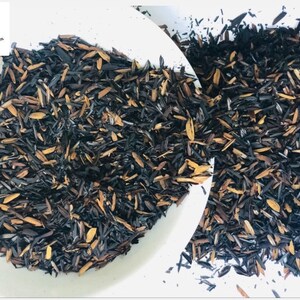 May include: A close-up of a bowl filled with loose black and brown tea leaves. The tea leaves are scattered on a white surface.