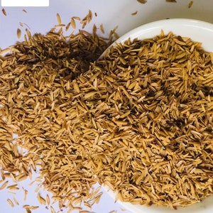 May include: A white bowl filled with brown rice husks, also known as rice bran, with more husks scattered on a white surface. The husks are a natural byproduct of rice production and are often used as a source of fiber and nutrients.