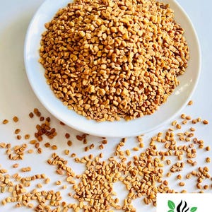 Puede incluir: Un plato blanco rebosante de semillas de alholva de color marr&oacute;n dorado, con algunas semillas esparcidas. La imagen est&aacute; bien iluminada, resaltando la textura y el color de las semillas. El logotipo de Ceylon Spices Foods est&aacute; en la esquina inferior derecha.