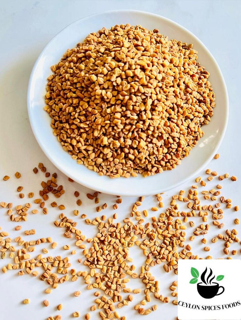 Puede incluir: Un plato blanco rebosante de semillas de alholva de color marr&oacute;n dorado, con algunas semillas esparcidas. La imagen est&aacute; bien iluminada, resaltando la textura y el color de las semillas. El logotipo de Ceylon Spices Foods est&aacute; en la esquina inferior derecha.