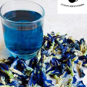 May include: A glass of blue butterfly pea flower tea with dried blue butterfly pea flowers scattered around the glass. The flowers are a vibrant blue with white centers and green stems. The text "CEYLON SPICES FOODS" is visible in the upper right corner of the image.