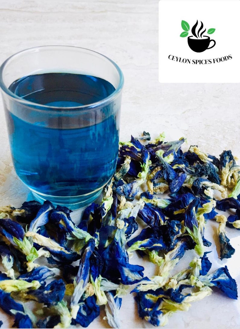 May include: A glass of blue butterfly pea flower tea with dried blue butterfly pea flowers scattered around the glass. The flowers are a vibrant blue with white centers and green stems. The text "CEYLON SPICES FOODS" is visible in the upper right corner of the image.