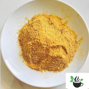 Puede incluir: Un plato blanco contiene un mont&oacute;n de polvo de especias amarillo-marr&oacute;n. El polvo tiene una textura fina y granulada. El logotipo de Ceylon Spices Foods est&aacute; en la esquina inferior derecha, con una taza negra con vapor y hojas verdes.