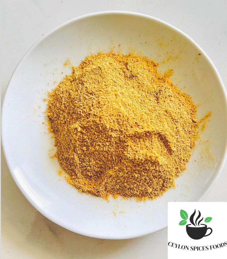 Puede incluir: Un plato blanco contiene un mont&oacute;n de polvo de especias amarillo-marr&oacute;n. El polvo tiene una textura fina y granulada. El logotipo de Ceylon Spices Foods est&aacute; en la esquina inferior derecha, con una taza negra con vapor y hojas verdes.