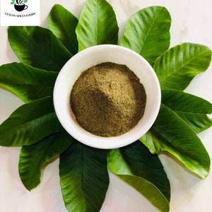 Puede incluir: Un tazón blanco lleno de polvo verde, rodeado de hojas verdes. El tazón está sobre una superficie blanca. El texto "CEYLON SPICES FOODS" es visible en la esquina superior izquierda de la imagen.