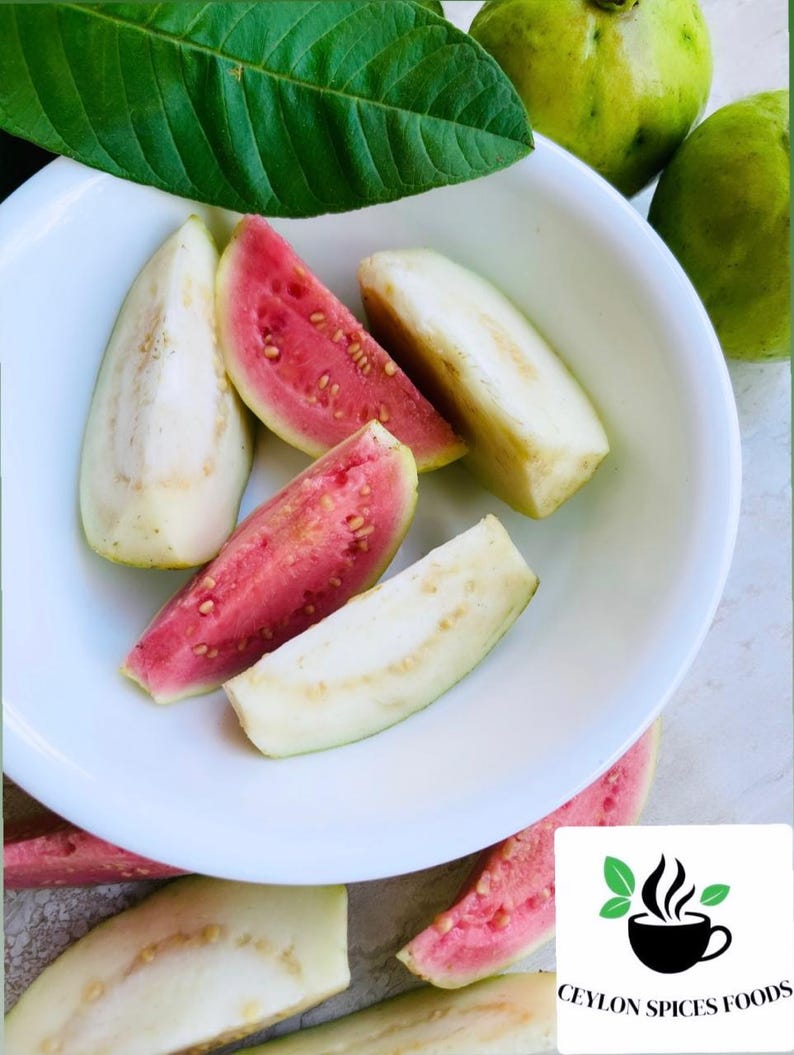 May include: A white bowl filled with sliced pink guava fruit. The bowl is on a table with a green leaf and whole guava fruit.