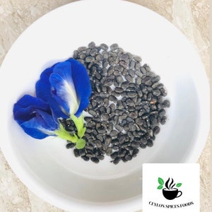May include: A white bowl with a pile of dark blue butterfly pea flower seeds and two blue butterfly pea flowers. The bowl is on a light brown surface. The text "CEYLON SPICES FOODS" is visible in the lower right corner of the image.