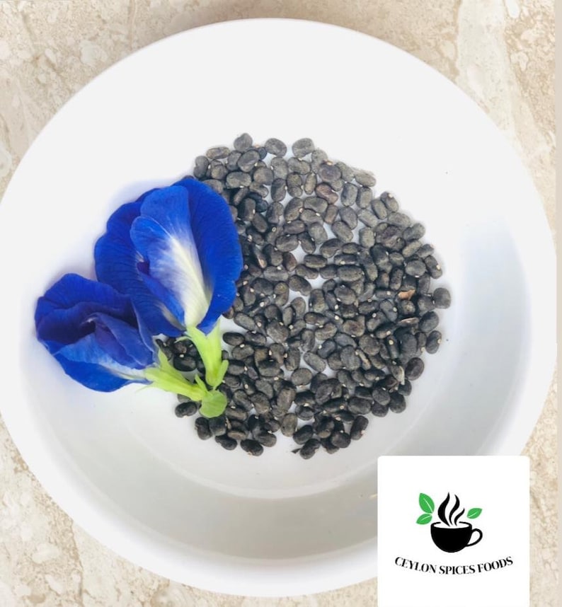 May include: A white bowl with a pile of dark blue butterfly pea flower seeds and two blue butterfly pea flowers. The bowl is on a light brown surface. The text "CEYLON SPICES FOODS" is visible in the lower right corner of the image.