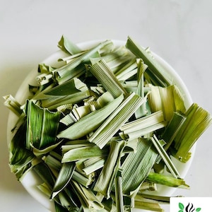 May include: A white bowl filled with dried, chopped lemongrass stalks. The pieces are light green and tan, featuring a fibrous, blade-like texture. A small logo in the corner displays a black coffee cup icon with green leaves and the text CEYLON SPICES FOODS.