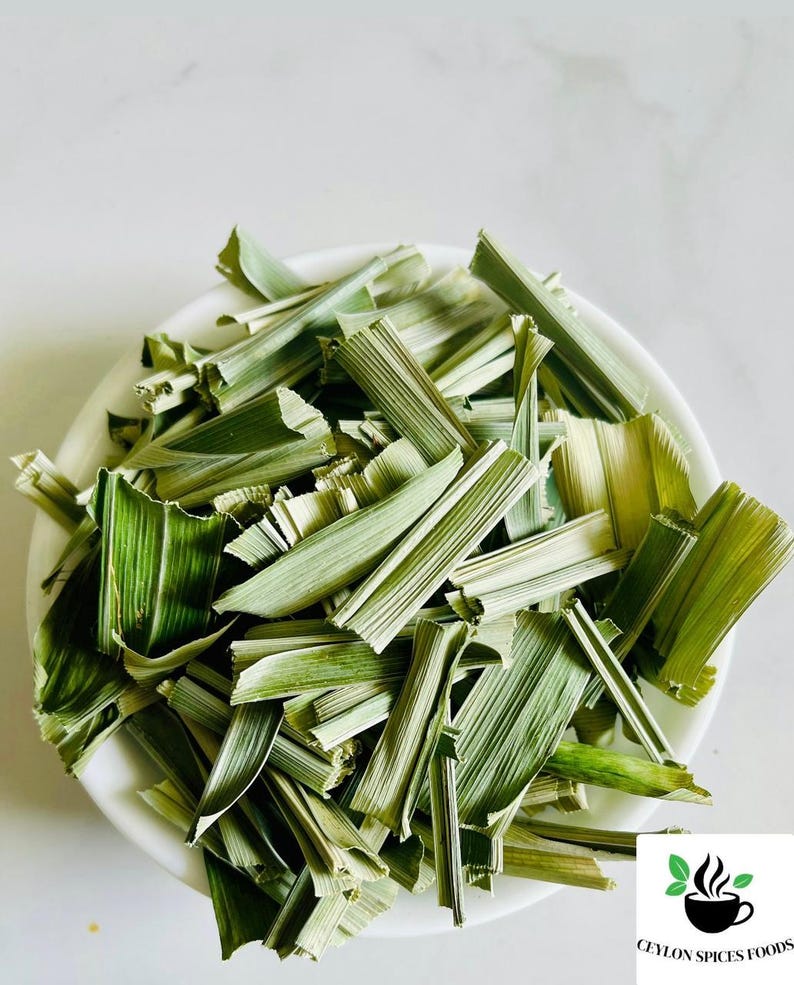 May include: A white bowl filled with dried, chopped lemongrass stalks. The pieces are light green and tan, featuring a fibrous, blade-like texture. A small logo in the corner displays a black coffee cup icon with green leaves and the text CEYLON SPICES FOODS.