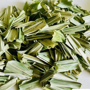 May include: A pile of dried, light green lemongrass stalks cut into small, uniform segments. The pieces are fibrous and have a pale, muted color. In the bottom right corner, a logo features a black coffee cup icon with green leaves.