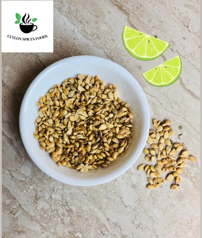 May include: A white bowl filled with dried, light brown watermelon seeds. A few seeds are scattered on the table next to the bowl. Two lime wedges are on the table.