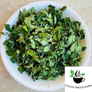 May include: A white bowl filled with dried green moringa leaves. In the bottom right corner, a white square logo features a black coffee cup icon with steam, two green leaves, and the black text CEYLON SPICES FOODS printed below the cup.