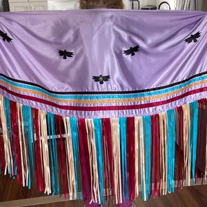 May include: A lilac-coloured shawl featuring black bird motifs and vibrant ribbon trim. The shawl has a fringe of red, blue, cream, and turquoise ribbons. The design is likely inspired by Native American traditions.