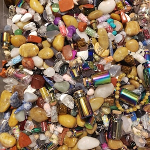 May include: A collection of polished gemstones and small glass bottles with iridescent metal caps. The stones vary in color, including yellow, brown, white, and red. Some bottles contain glitter or small objects, suggesting a craft or jewelry theme.