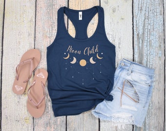 May include: A navy blue tank top with the text "Moon Child" and a graphic of a crescent moon with stars. The tank top is lying on a light wood surface with a pair of pink flip flops and a pair of light blue denim shorts.