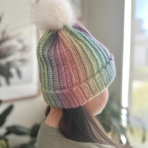 Può includere: Un berretto lavorato a maglia a righe nei colori pastello arcobaleno, rosa, blu, verde e viola. Ha un design a coste e un soffice pompon bianco. Un accessorio invernale caldo e alla moda.