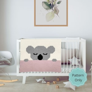 May include: A crochet baby blanket featuring a grey koala design. The blanket is white with a pink border. The blanket is on a white cot in a nursery.
