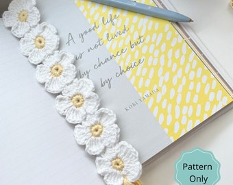 May include: A white crocheted bookmark with a chain of daisy flowers and a yellow tassel. The bookmark is on a yellow and white patterned journal with the text "A good life is not lived by chance but by choice" and "KOBI YAMADA". The text "Pattern Only" is on a teal circle.
