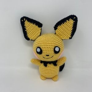 May include: A handmade crochet Pikachu doll. The doll is primarily yellow with black accents on the ears and tail. It has black eyes, a small smile, and pink cheeks. The arms and legs are small and rounded, and the doll is standing upright.